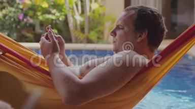 胖懒人光着胸膛躺在吊床上用手机在游泳池边看书。 3840x2160